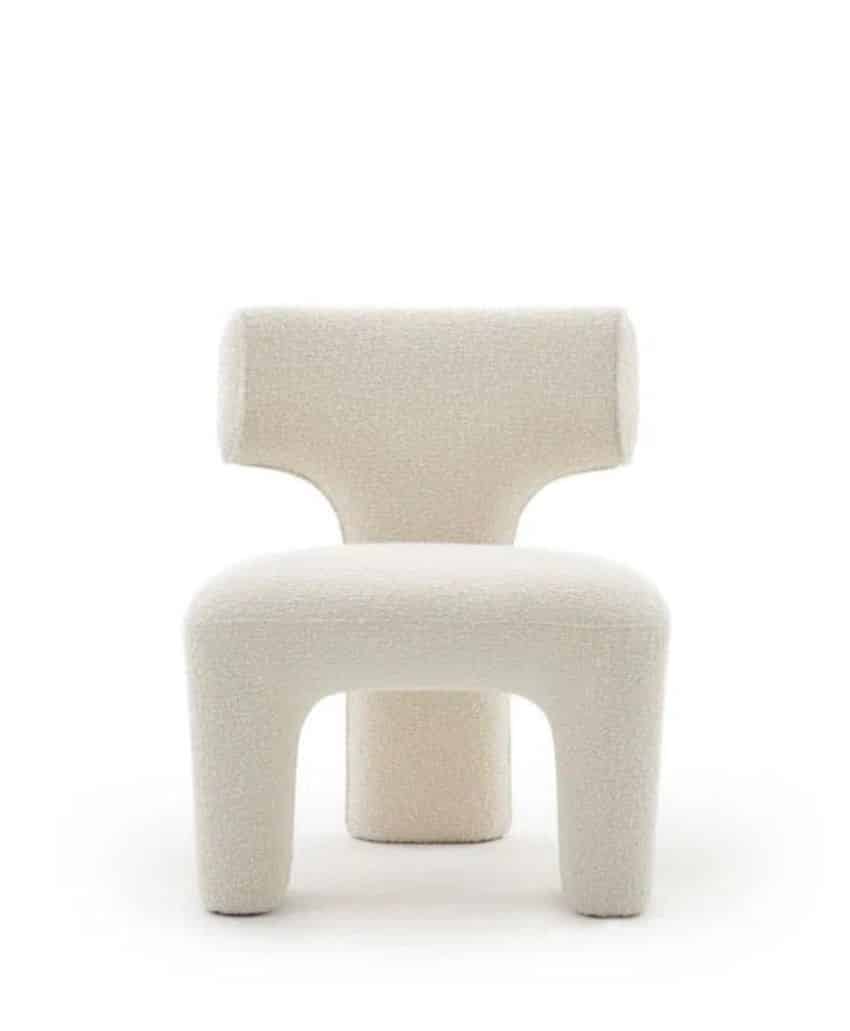 A modern, minimalist chair with three thick, rounded legs and a wide, T-shaped backrest, upholstered in textured off-white fabric, against a plain white background.