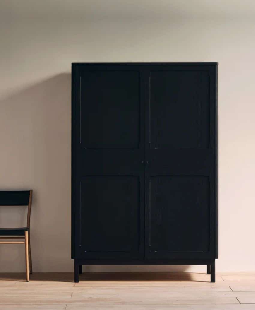 A tall, black, four-door cabinet stands against a light beige wall next to a wooden chair with a black seat and backrest. The floor is light wood.