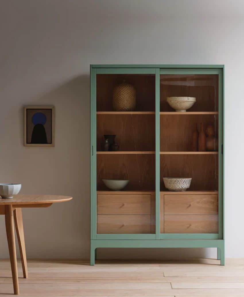 A minimalistic interior featuring a light green-framed glass cabinet with wooden shelves, displaying bowls and decorative objects. A small wooden table and an abstract artwork hang on the light-colored wall beside it.