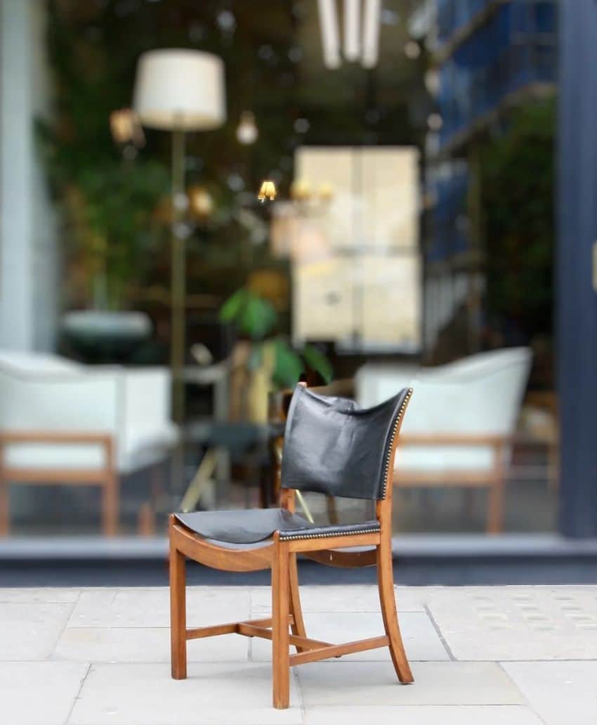 A wooden chair with black leather upholstery sits on a sidewalk in front of a glass window, through which modern indoor furniture and decor, including lamps and plants, are visible.