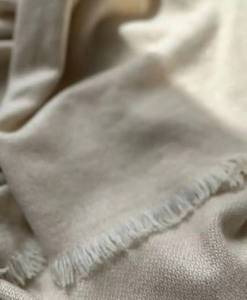 A close-up of a soft, cream-colored fabric with fringed edges, slightly wrinkled and folded, creating a cozy and textured appearance.