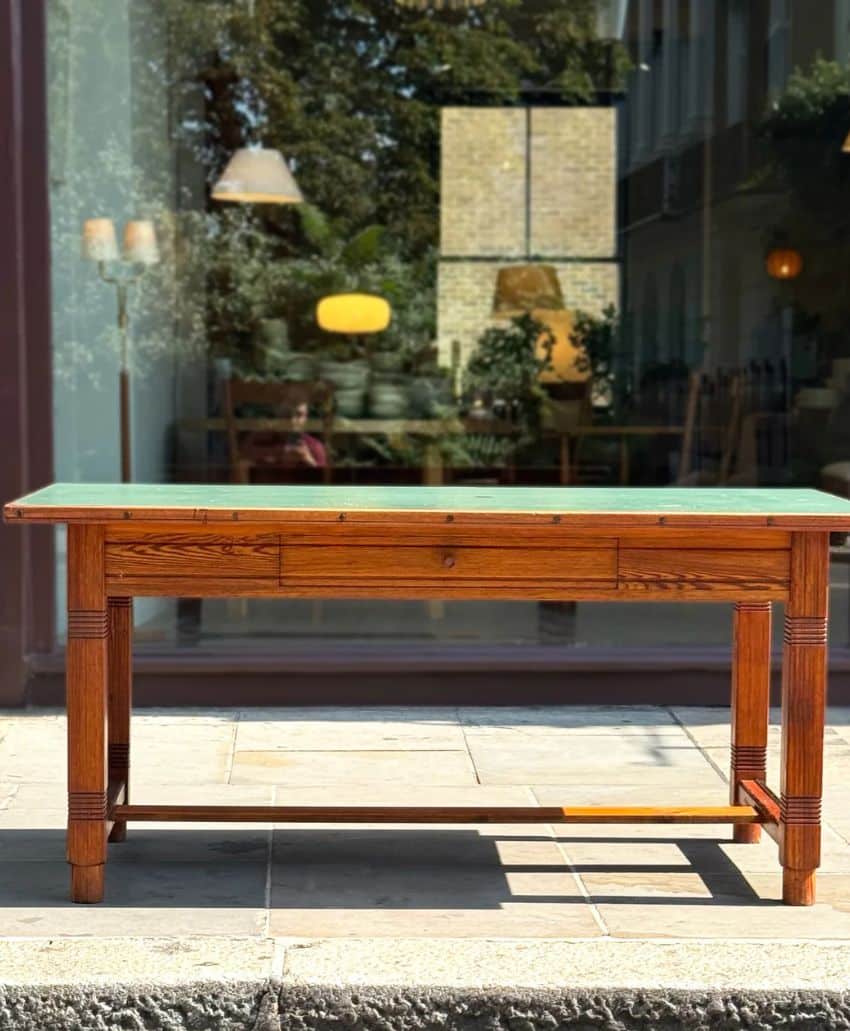 A wooden table with a green top stands on a stone patio in front of a large glass window reflecting outdoor trees and indoor lamps.