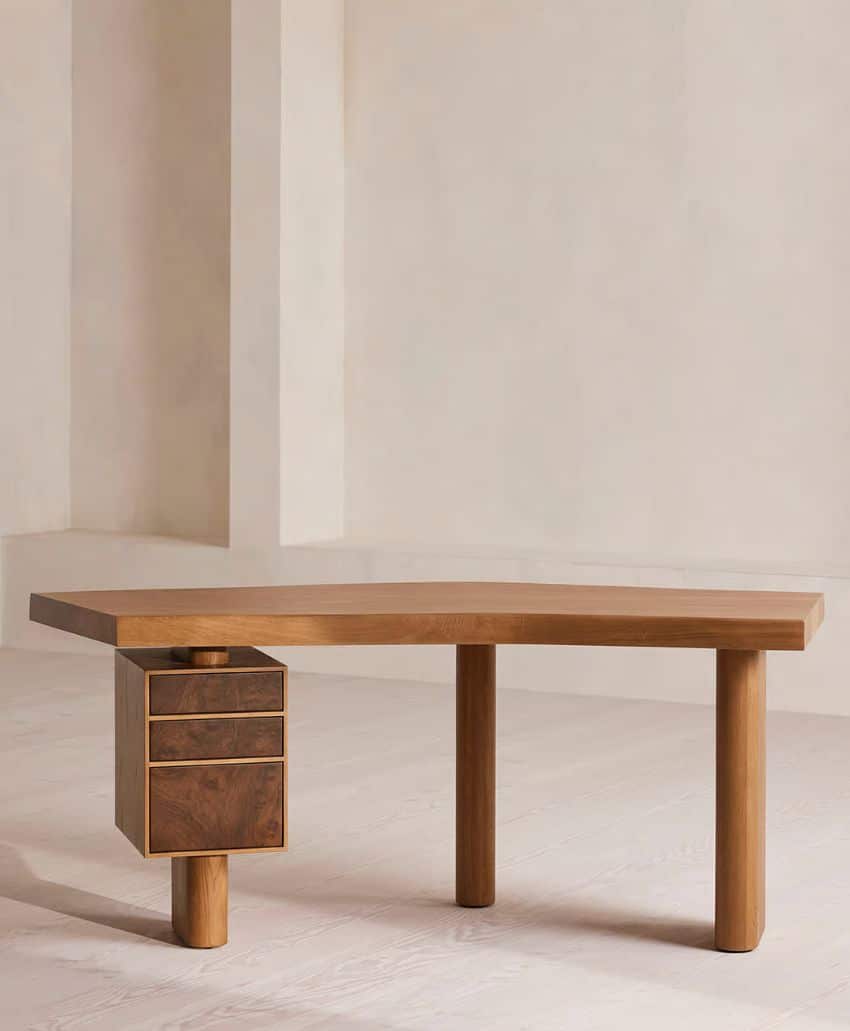 A modern wooden desk with an angled top, two cylindrical legs on the right, and a set of three small drawers on the left supported by a short round leg, set against a neutral background.