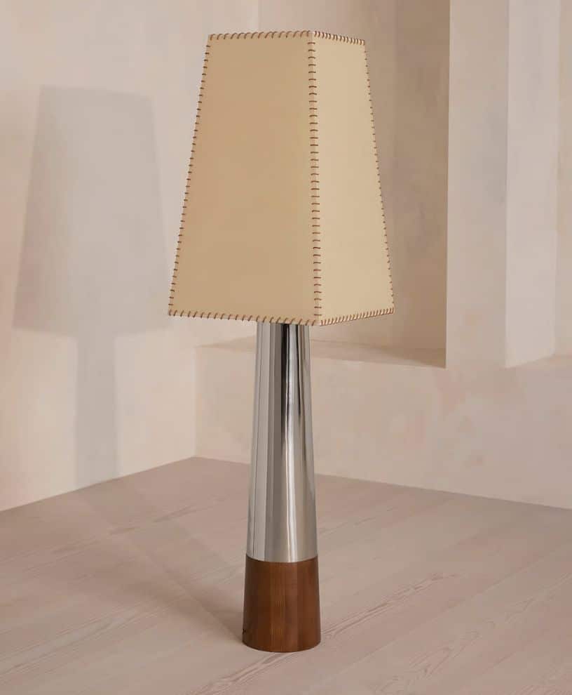 A modern floor lamp with a tapered beige lampshade, stitched along the edges, sits on a shiny silver and wooden base against a light-colored wall and floor.