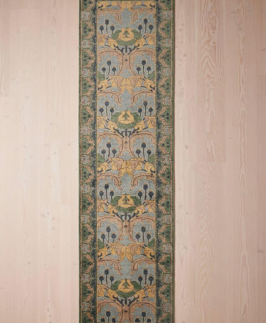 A long, narrow rug with an intricate floral pattern in shades of green, gold, and blue, bordered by a darker green edge, displayed on a pale wooden floor.