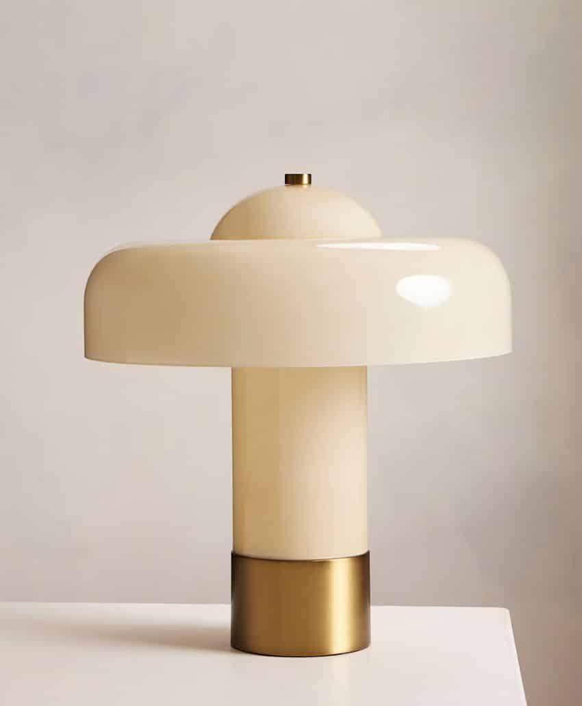 A modern, cream-colored table lamp with a round, glossy shade and brass base sits on a white surface against a plain, light-colored background.