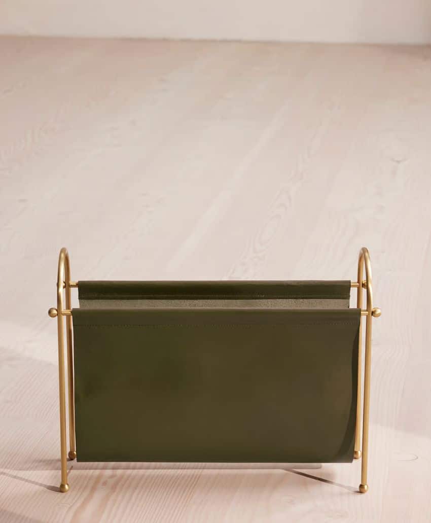 A modern magazine rack with a green leather sling and gold metal frame sits on a light wood floor in a minimalist setting.