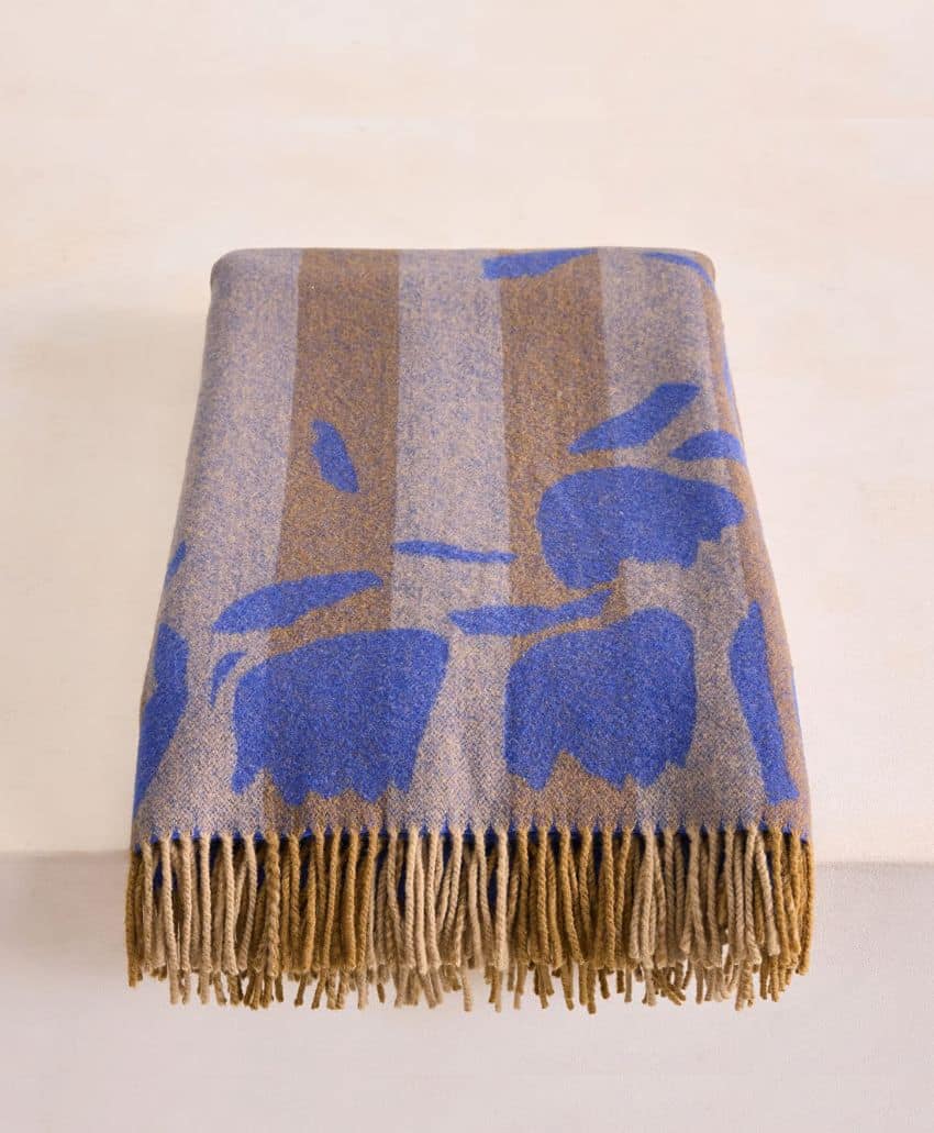 A folded wool blanket with brown and gray stripes, blue abstract floral patterns, and brown fringe on the bottom edge, placed on a light surface.