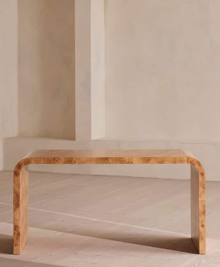 A minimalist, rectangular wooden table with rounded edges sits on a light-colored wooden floor against a neutral, textured wall background. The table has a smooth, natural wood finish.