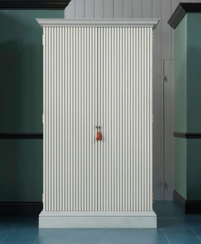 A tall, white, vertically ribbed cabinet with gold hinges and a small red tassel hanging from the handles, standing against a two-tone green and gray paneled wall.
