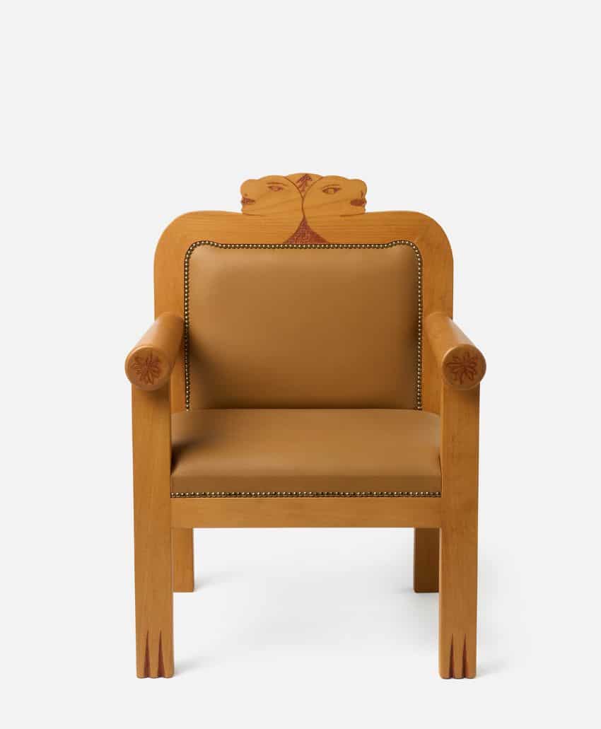 A wooden armchair with tan upholstery, decorative carvings on the arms and backrest, and a unique double-face motif carved at the top of the backrest.