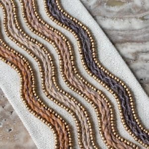 A close-up of decorative trim featuring wavy braided threads in earth tones, bordered by rows of small gold beads, on a neutral fabric background atop a marble surface.