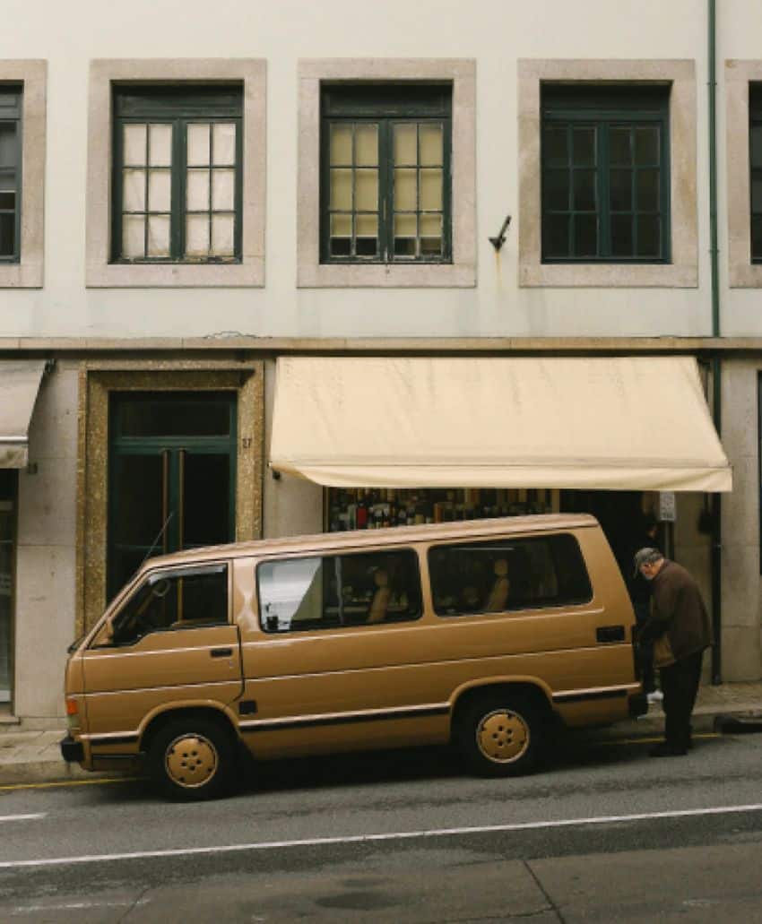 A beige van is parked in front of a building with large windows and a yellow awning. An elderly person stands by the van’s rear door on a city street.