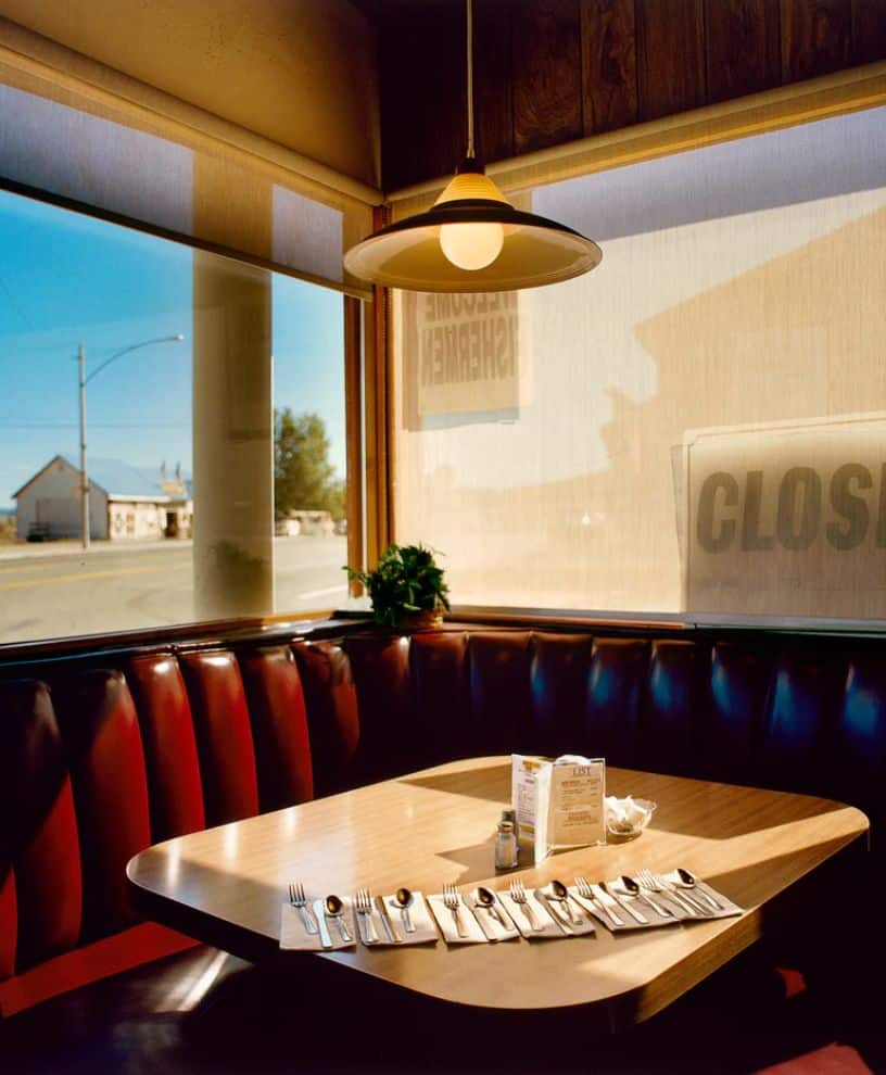 A sunlit diner booth with red vinyl seats and a wooden table set with utensils, napkins, and condiments. The window shows a quiet street outside and a sign that reads CLOSED.