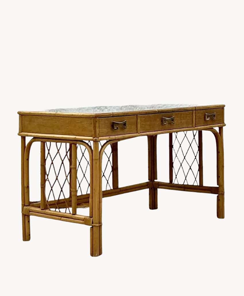 A rectangular wooden desk with three drawers and a lower lattice shelf stands on a concrete surface in front of a textured, weathered gray wall.