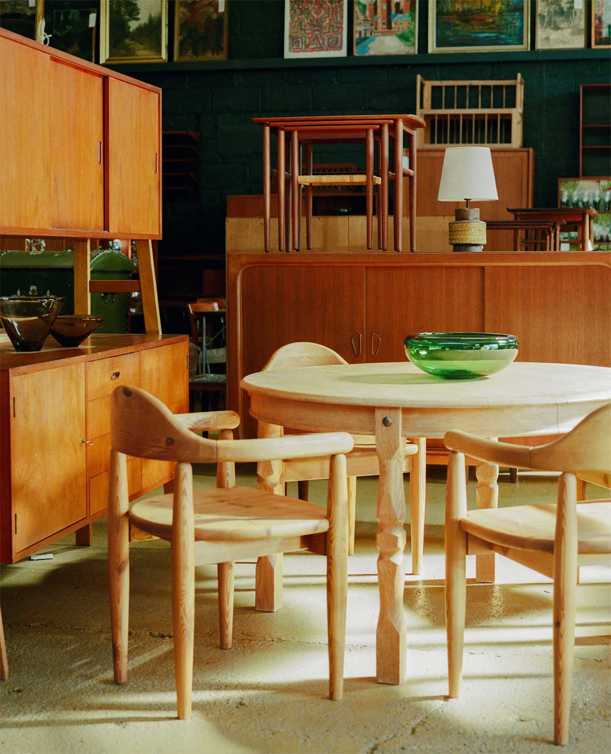 A vintage-style room with wooden furniture, including a round table, four chairs, cabinets, a green glass bowl on the table, and framed pictures on a dark wall in the background.