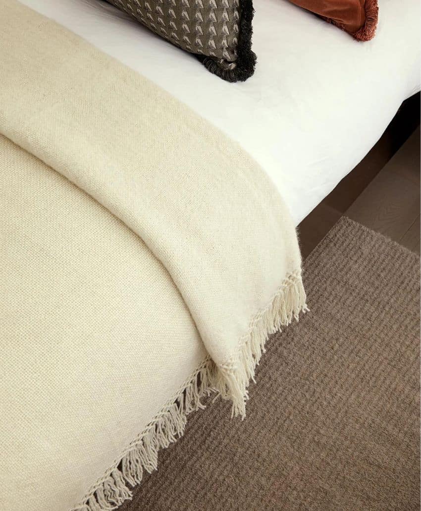 A cream-colored fringed throw blanket is draped neatly over the edge of a bed with white bedding. Decorative pillows in dark and rust colors are arranged at the head of the bed, and a textured beige rug is on the floor.