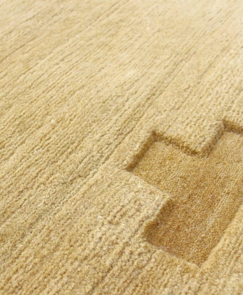 A close-up of a beige textured carpet with a section cut out in the shape of a plus sign, revealing a slightly lower layer beneath.