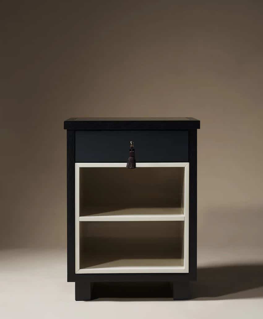 A small, dark wooden nightstand with a single drawer featuring a tassel handle and two open cream-colored shelves below, set against a neutral background.