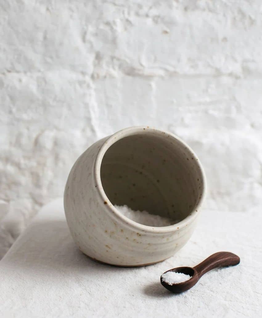 A ceramic bowl filled with coarse salt lies on its side, with some salt spilled out onto a white surface. A small wooden spoon with salt rests nearby. The background is a textured white wall.