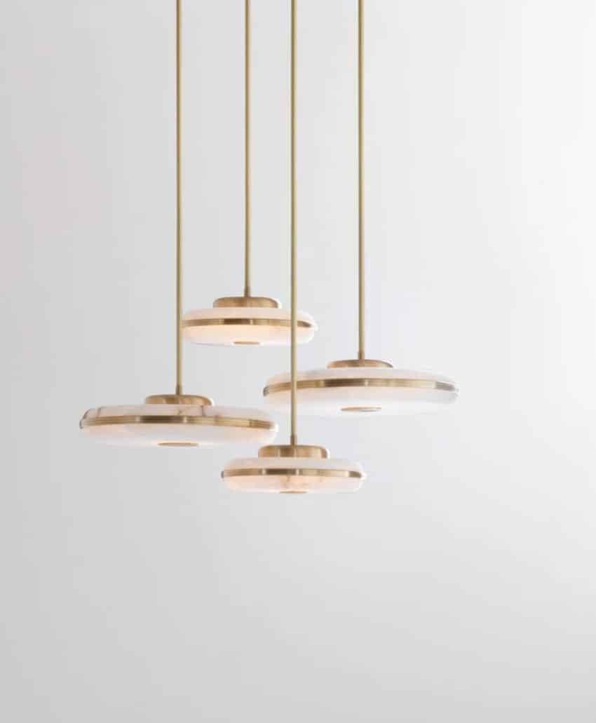 Four modern pendant lights with round, flat, golden fixtures hang at different heights against a plain, light grey background. The minimalist design creates a clean and elegant appearance.