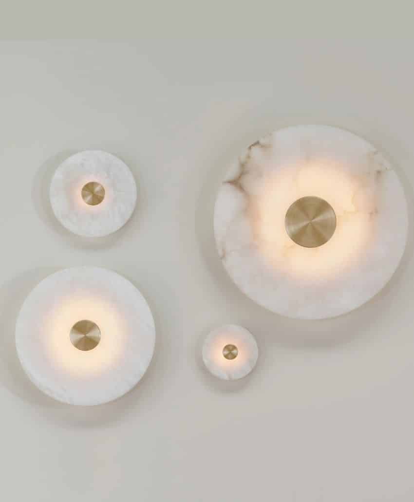 Four round wall lights with brass centers and diffused, glowing white edges are mounted on a light wall in an asymmetric arrangement. The lights vary in size, creating a modern, artistic display.