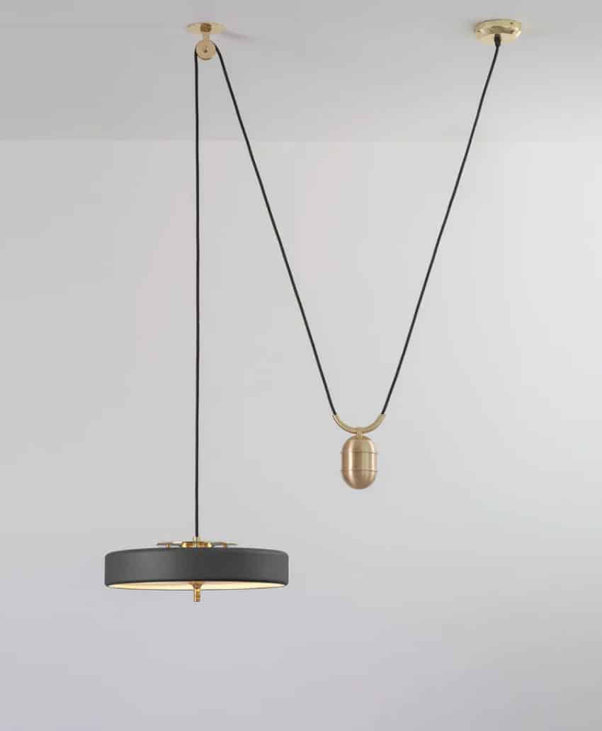 A modern pendant light with a black lampshade hangs from the ceiling, suspended by a black cord looped through two brass ceiling mounts, creating a V-shape with a brass weight. The fixture contrasts against a white ceiling.