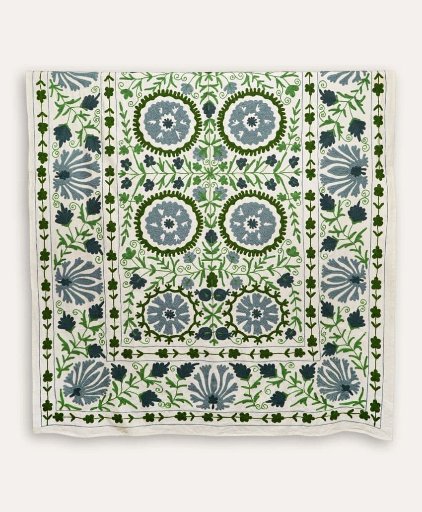 A rectangular textile featuring an intricate floral pattern in shades of green and blue on a white background, with symmetrical circular and leafy designs bordered by vines.