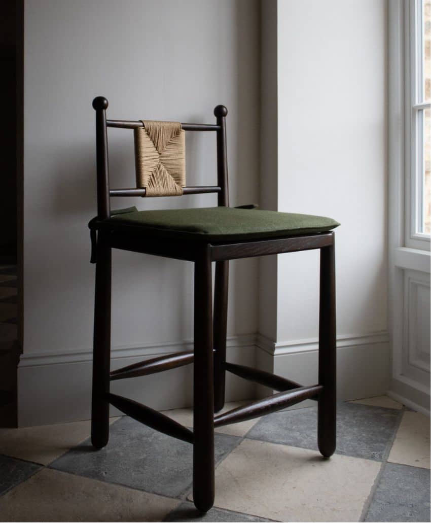 A dark wooden bar stool with a woven backrest and green seat cushion stands on a checkered tile floor near a window in a softly lit room.
