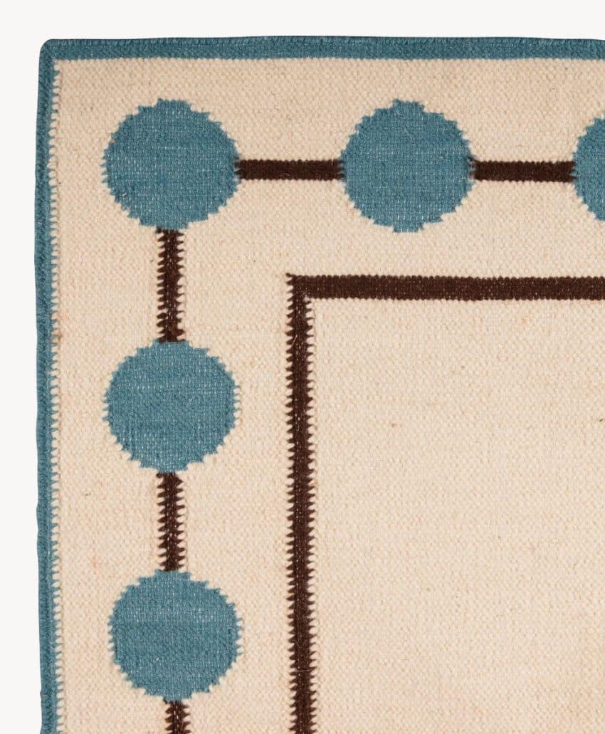 A section of a beige rug with a blue border decorated with blue circles connected by brown lines; the inner edge also has a thin brown border.