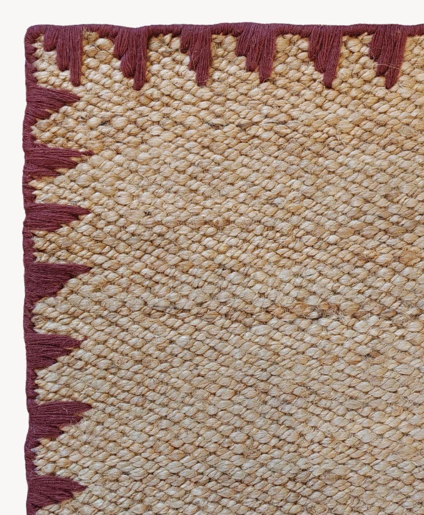A close-up of a woven natural fiber rug with a textured pattern, featuring a border of maroon triangular shapes along the edge.
