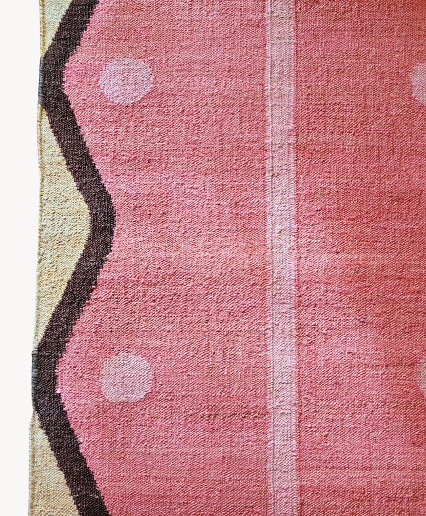 A close-up of a textured woven fabric with a pink, black, and tan abstract geometric pattern and white dots, shown against a white background.