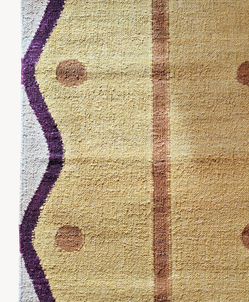 A close-up of a woven rug with a tan and yellow background, featuring a central brown vertical stripe, brown circles, and a wavy purple border along the left edge.