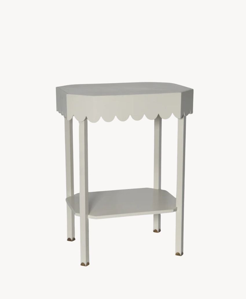 A small white side table with scalloped edges, four straight legs, and a lower shelf. The table has a simple, modern design with gold-colored tips on the legs.
