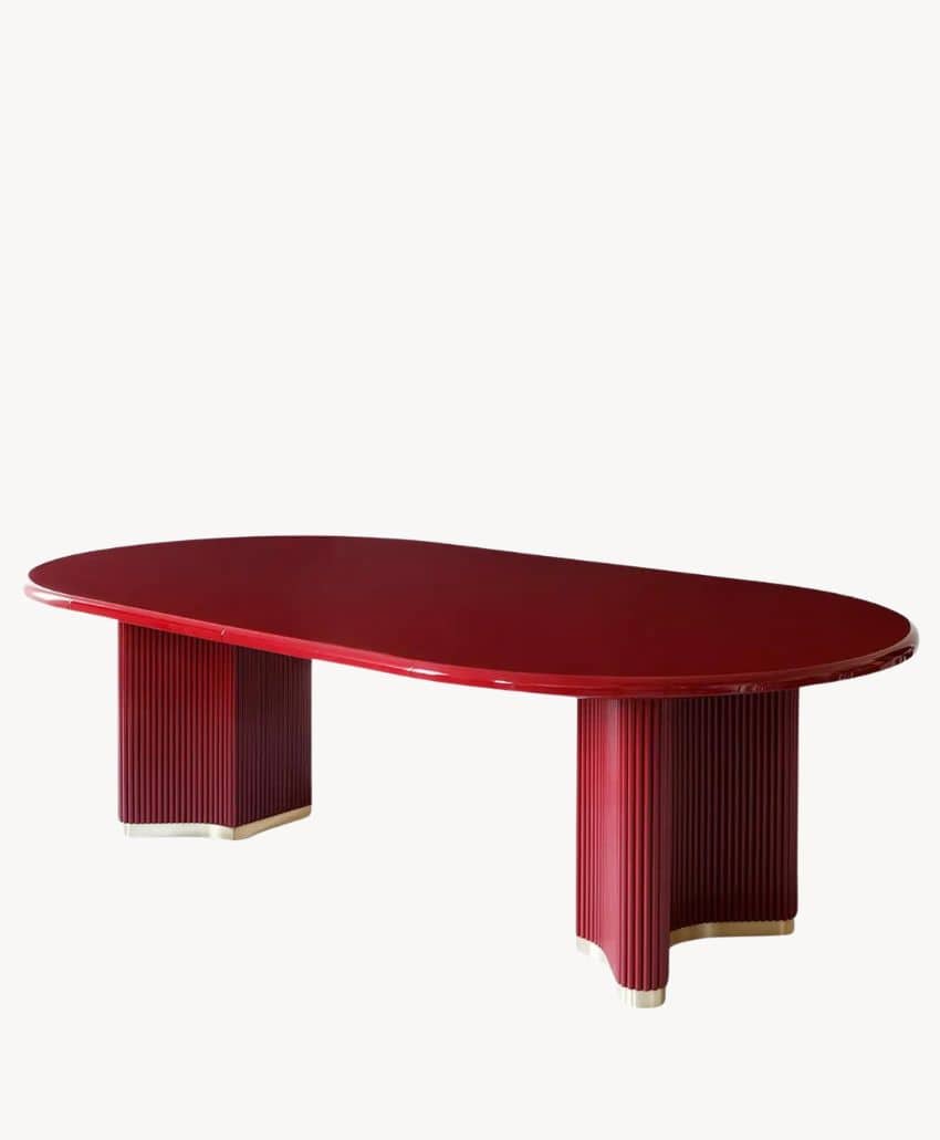 A modern, oval-shaped red table with a glossy finish, supported by two wide, vertically grooved pedestal legs. The table has a sleek, contemporary design against a plain white background.