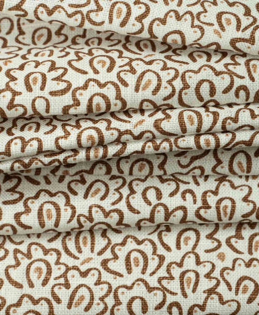 Beige fabric with a repeating, abstract brown floral pattern, shown in several folded layers. The texture of the material and the intricate design are clearly visible.