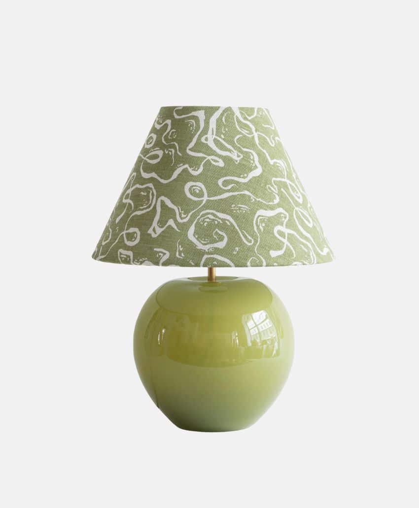 A table lamp with a round, glossy light green base and a green lampshade featuring a white abstract squiggle pattern, set against a plain white background.