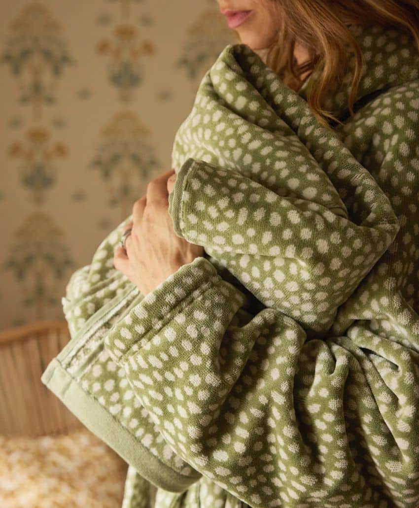 A person wearing a green and white patterned robe holds a baby wrapped in a matching blanket, standing in a softly lit room with patterned wallpaper in the background.