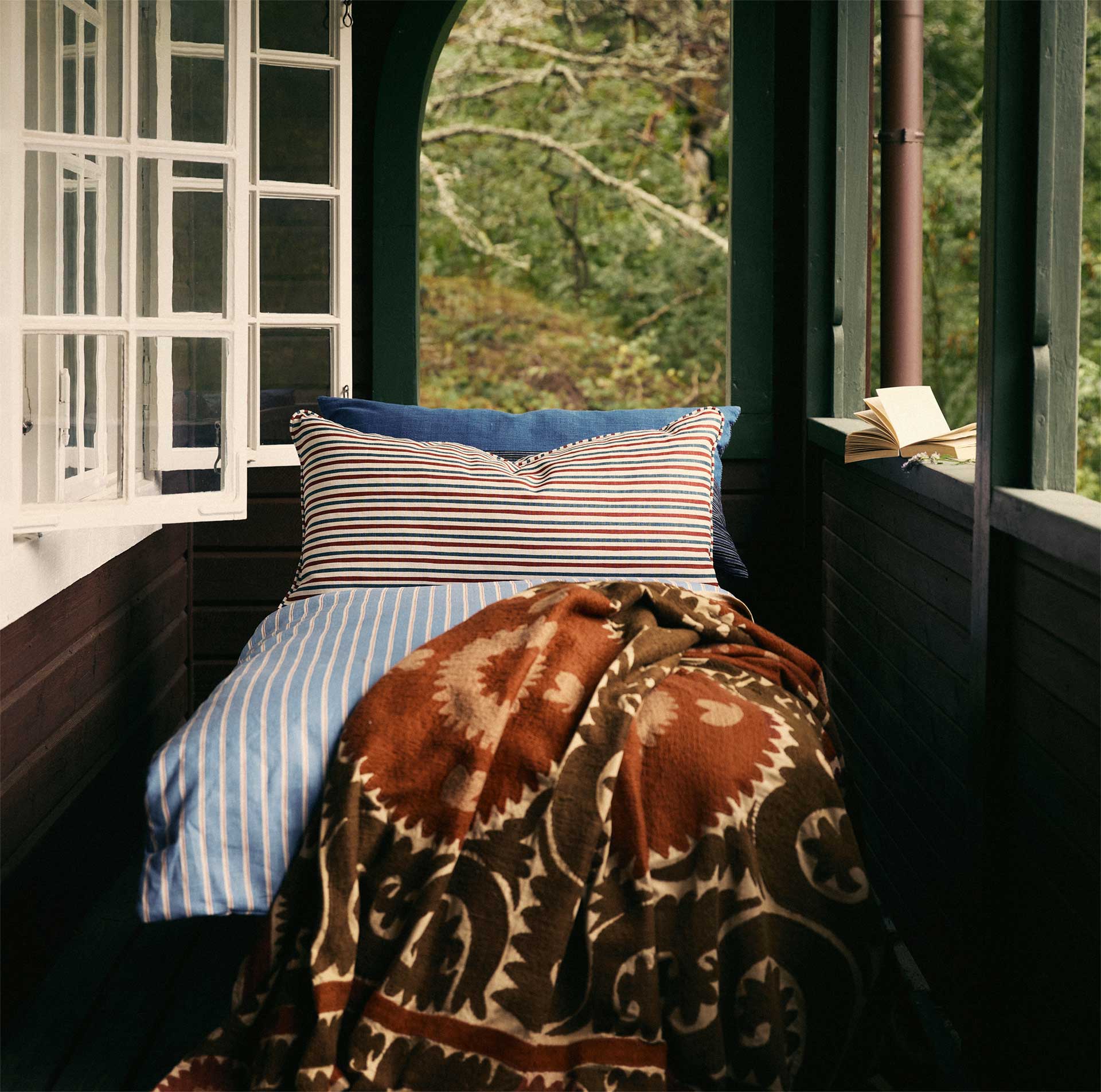 A cozy bed with striped pillows and a patterned blanket is placed next to open windows, letting in natural light with a view of green trees outside. An open book rests on the wooden window ledge.