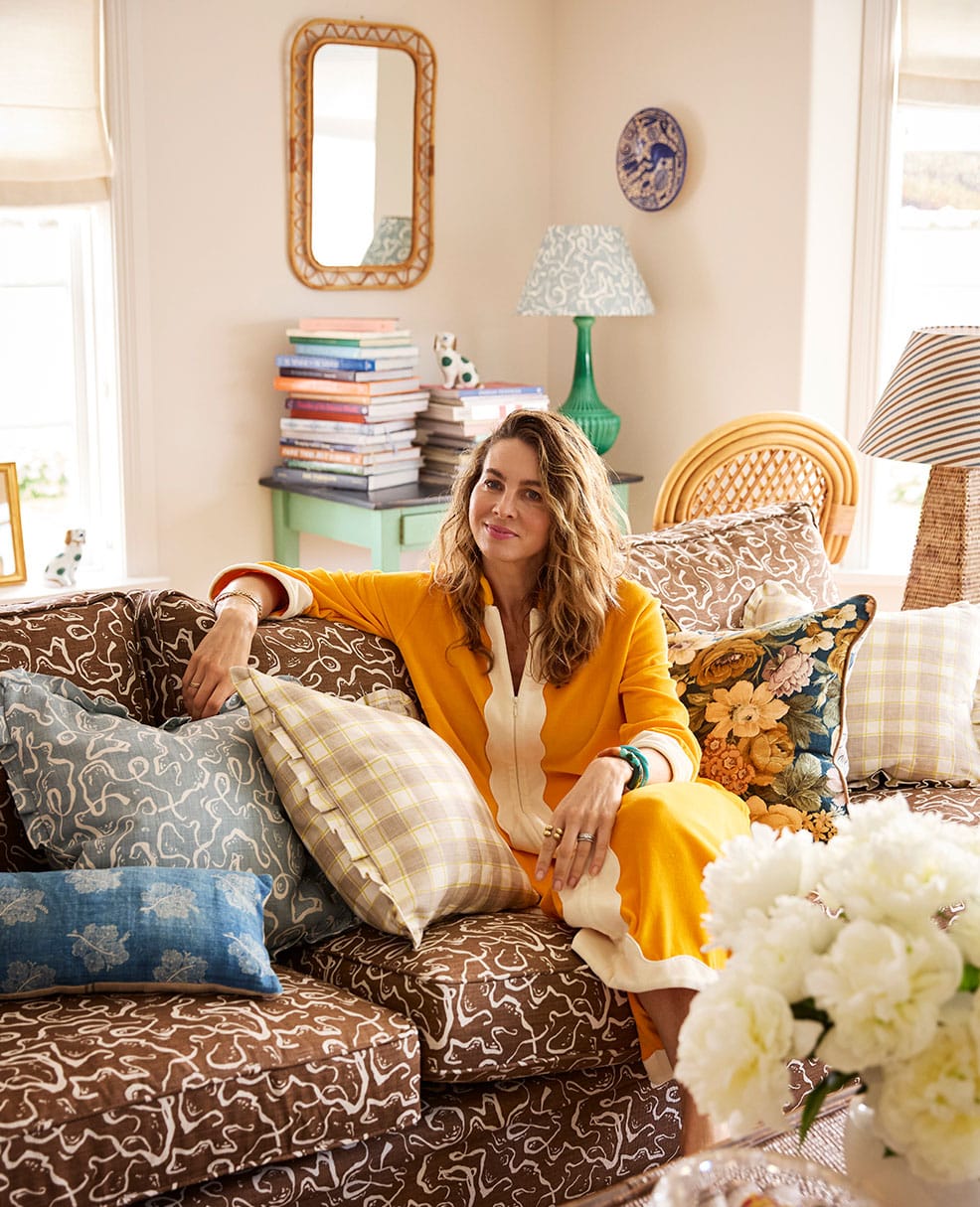 A woman in a yellow dress sits on a patterned brown sofa with various colorful pillows. Behind her are stacked books, a green lamp, a mirror, and bright decor in a cozy, sunlit living room. White flowers are in the foreground.