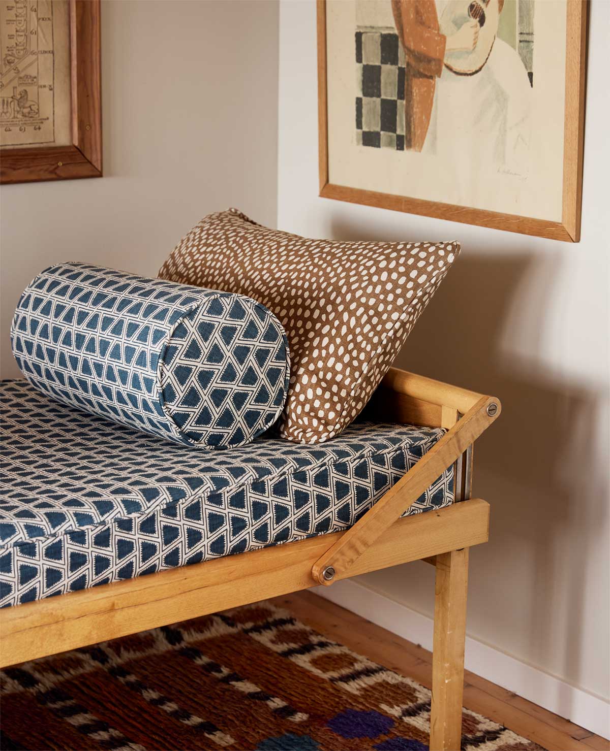 A wooden daybed with a blue geometric-patterned mattress, a brown-and-white dotted pillow, and a blue patterned bolster sits on a colorful rug next to framed artwork on a light-colored wall.