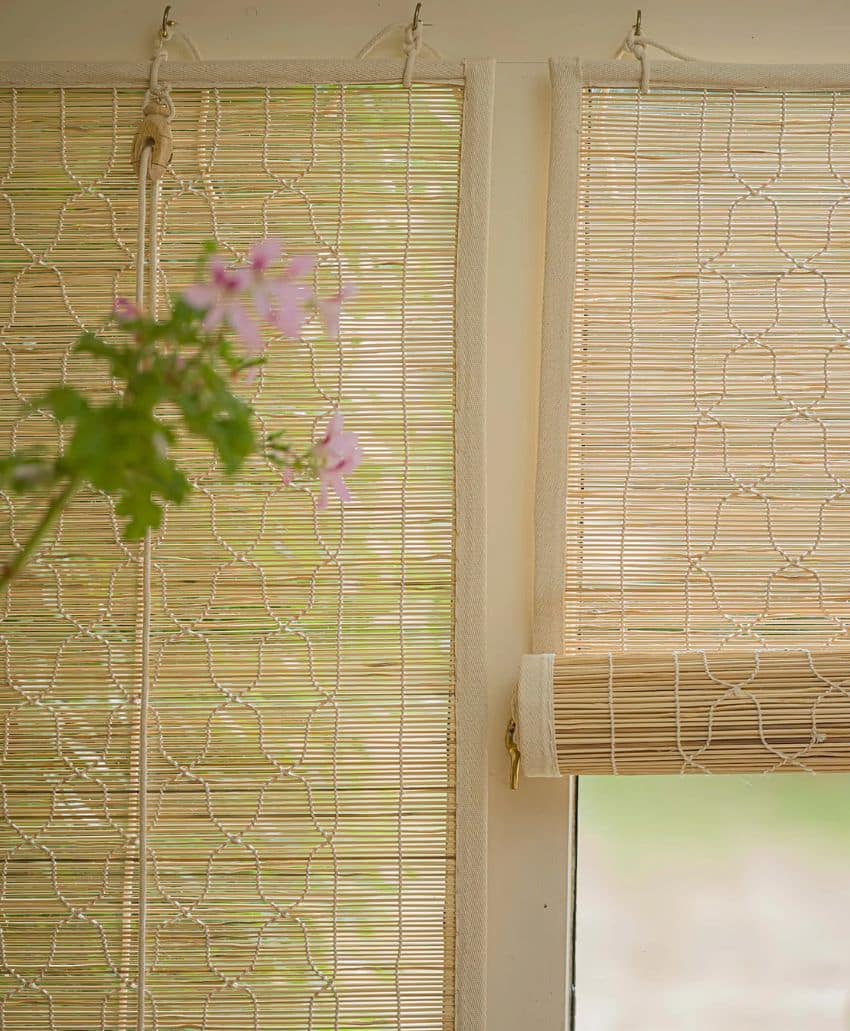 Two light bamboo blinds hang on a window, one partly rolled up. A blurred pink flower with green leaves is in the foreground, and soft natural light filters through the blinds.
