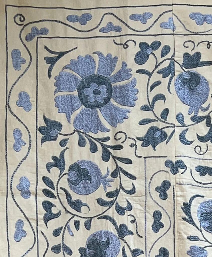 Embroidered fabric featuring a floral design in shades of blue and gray on a beige background, with large flowers, leaves, and swirling vines bordered by a decorative edge.