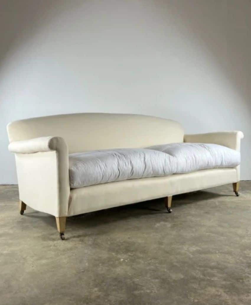 A cream-colored, vintage-style sofa with a curved backrest, plush light gray seat cushion, and wooden legs on small wheels, set against a plain wall and concrete floor.