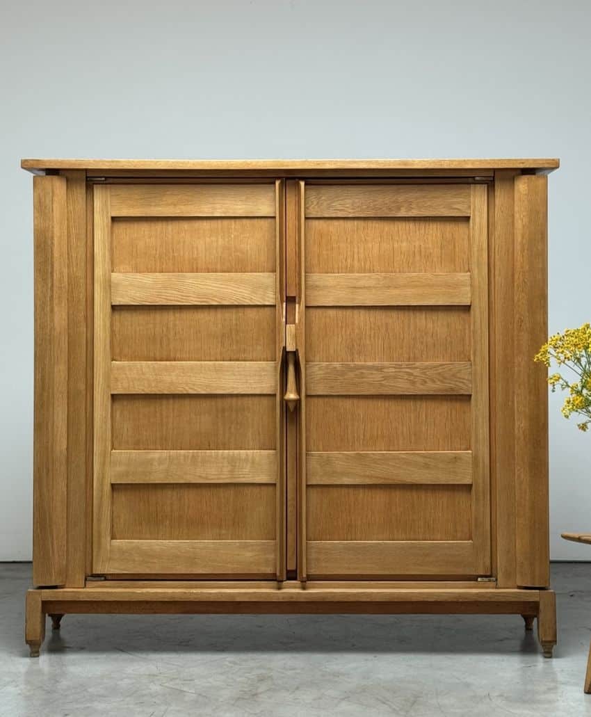A wooden cabinet with two vertical doors and horizontal panel details, standing on short legs. The cabinet has a natural wood finish and a simple, minimalist design. A small yellow flower arrangement is partially visible on the right.