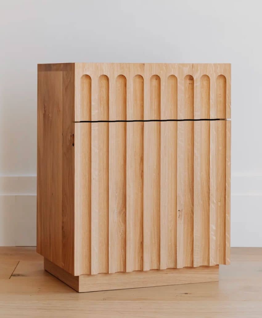 A small wooden cabinet with a natural finish and vertical grooves on the front, standing on a light wooden floor against a white wall.