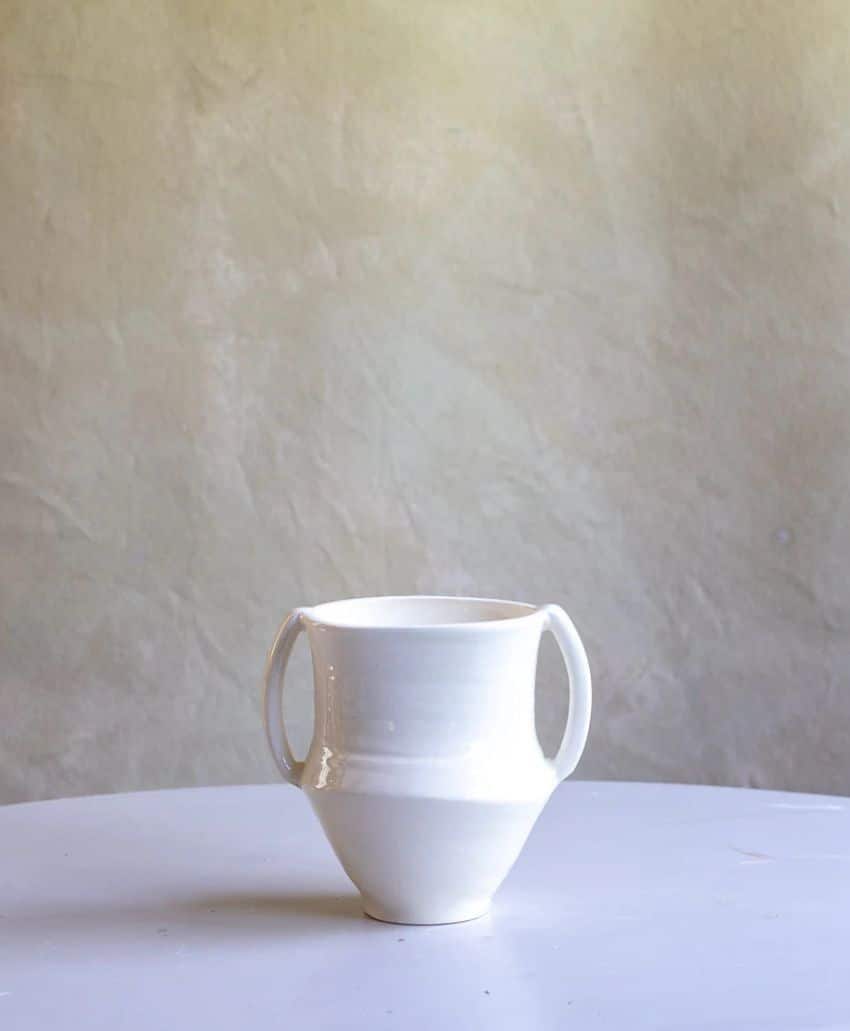 A white ceramic cup with three handles sits on a round white surface, against a textured beige background.