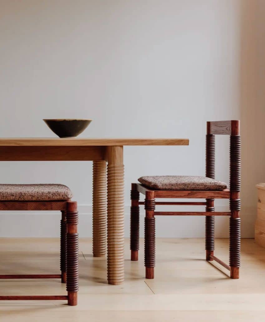 A minimalist room with a wooden table and two matching chairs featuring ribbed legs and woven seats; a single dark bowl sits on the table. The background is a plain white wall and light wood floor.