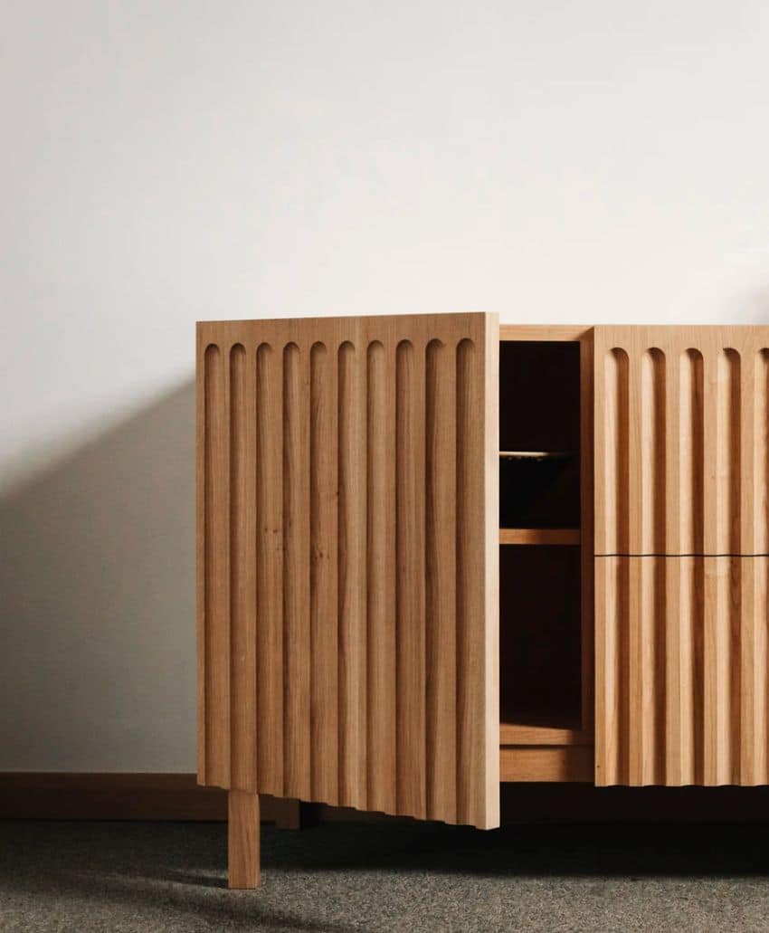 A wooden cabinet with vertical grooves on its doors, standing on short legs, with one door slightly open, revealing a dark interior against a plain white wall and carpeted floor.