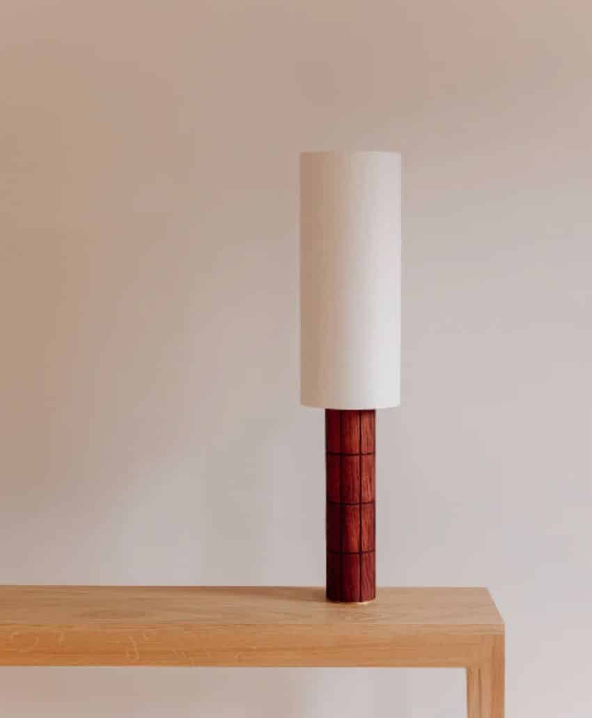 A modern table lamp with a cylindrical white shade and a wooden, square-patterned base sits on a light wooden table against a plain beige wall.